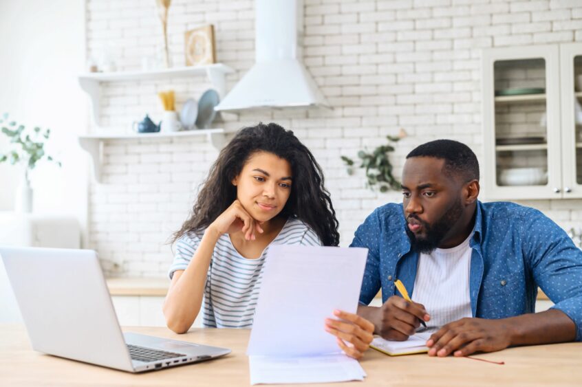 Mixed-race woman looking at paper with a laptop near her. A black man is seated next to her with a pen in his hand and a notebook looking at the sheet of paper that the mixed-race woman has.